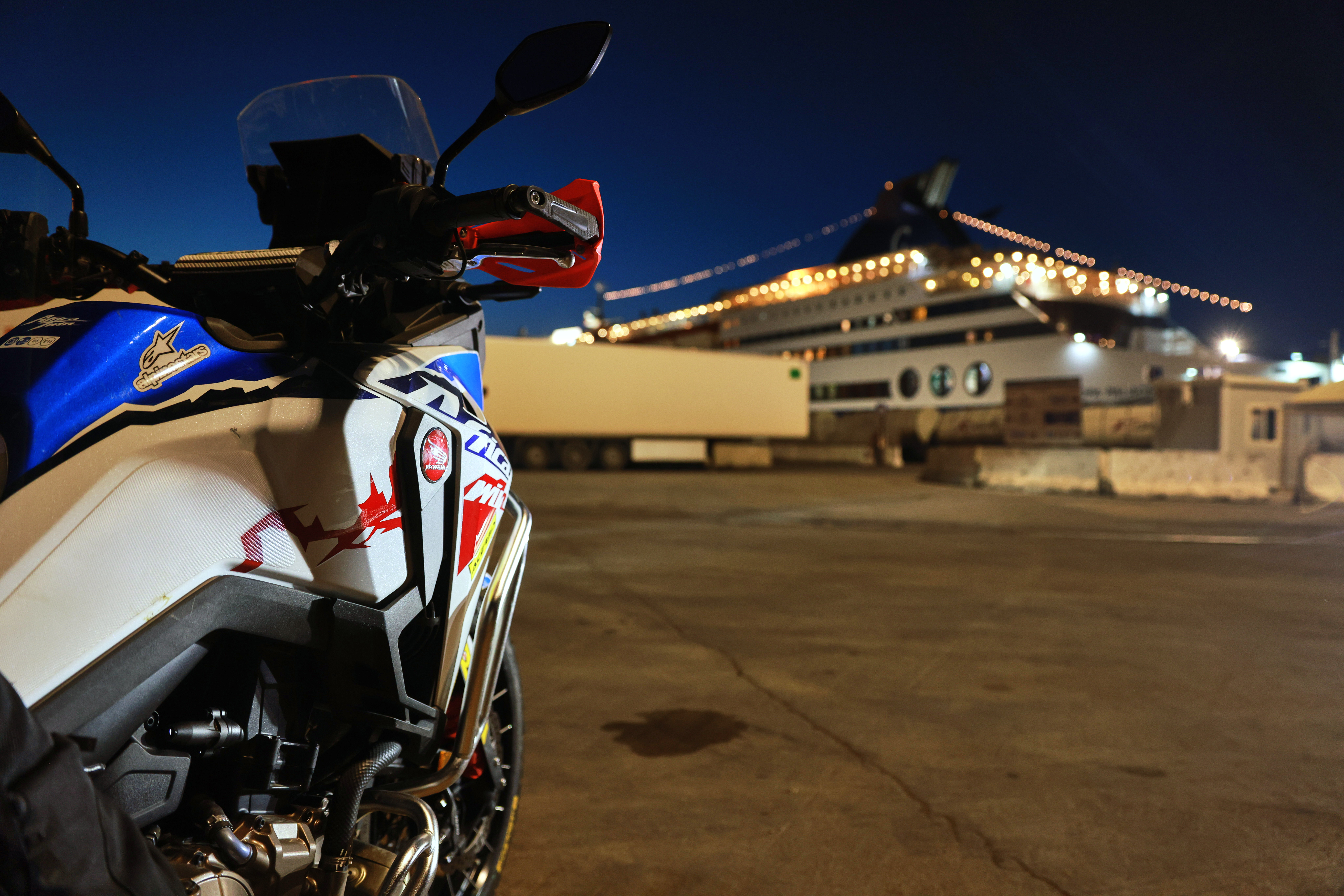 Una moto Honda Africa Twin in un porto, con una nave da crociera attraccata sullo sfondo di notte.